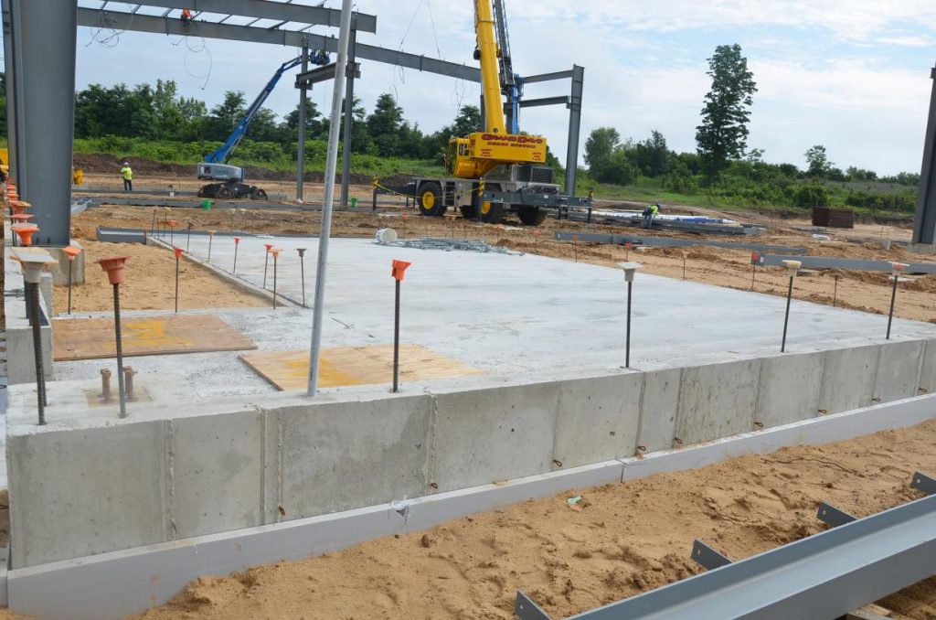 Construction site with concrete foundation and steel beams. Cranes and workers are visible in the background.