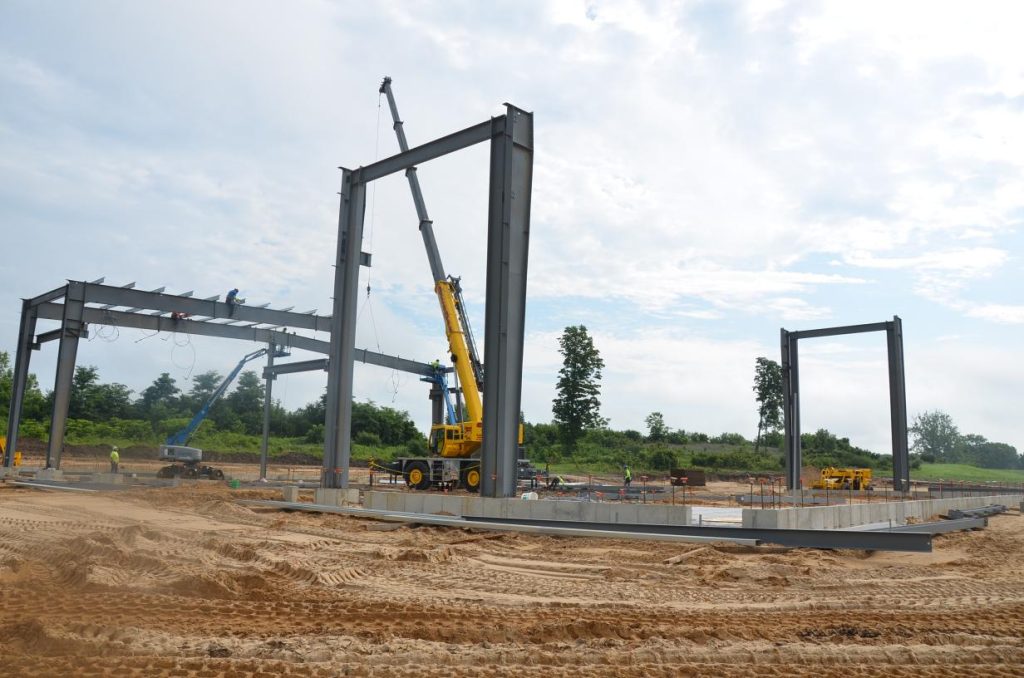 Construction site with steel framework partially erected, a yellow crane, and workers operating machinery; a grassy area is visible in the background under a cloudy sky.