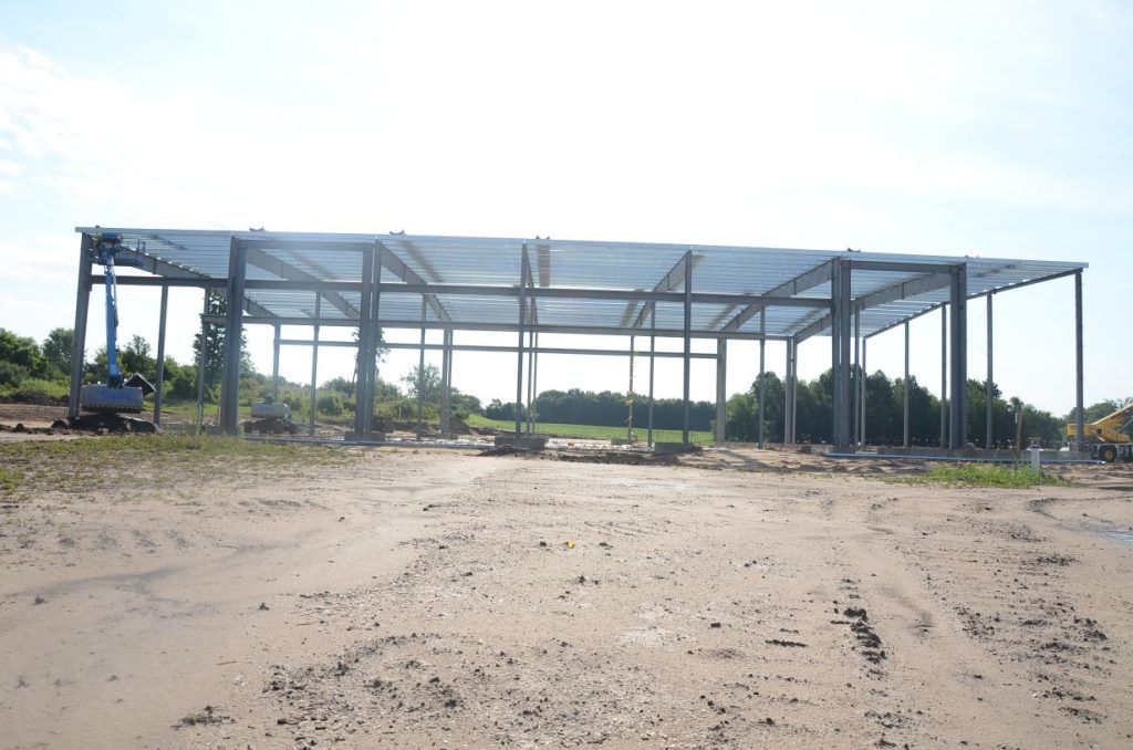 Steel framework of a large building under construction on a dirt lot, with trees and machinery in the background.