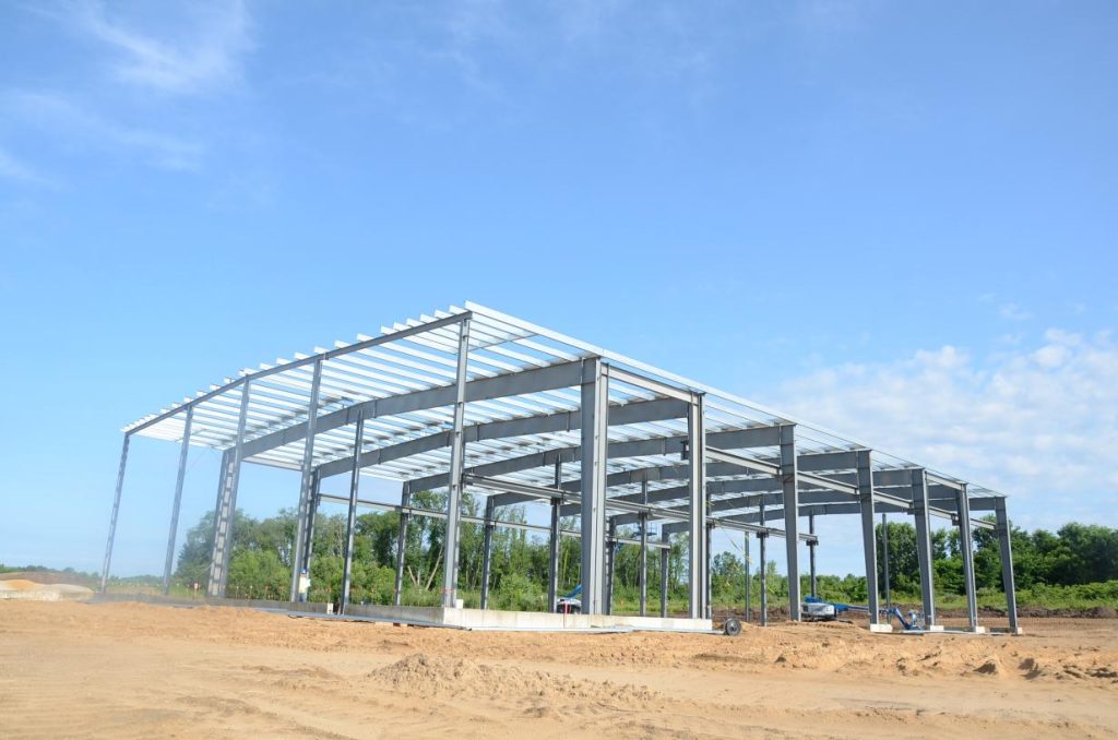Metal framework of a large building under construction on a sandy site with a clear blue sky and trees in the background.