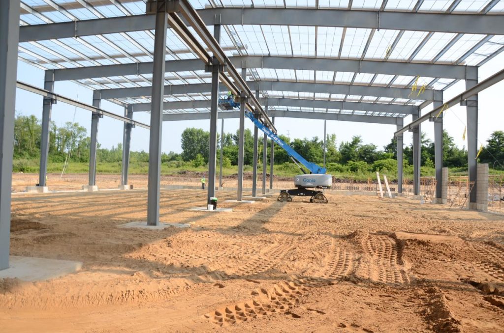 Construction site with a large metal framework and a blue boom lift under a partially completed roof. The ground is covered in dirt, and there are trees in the background.