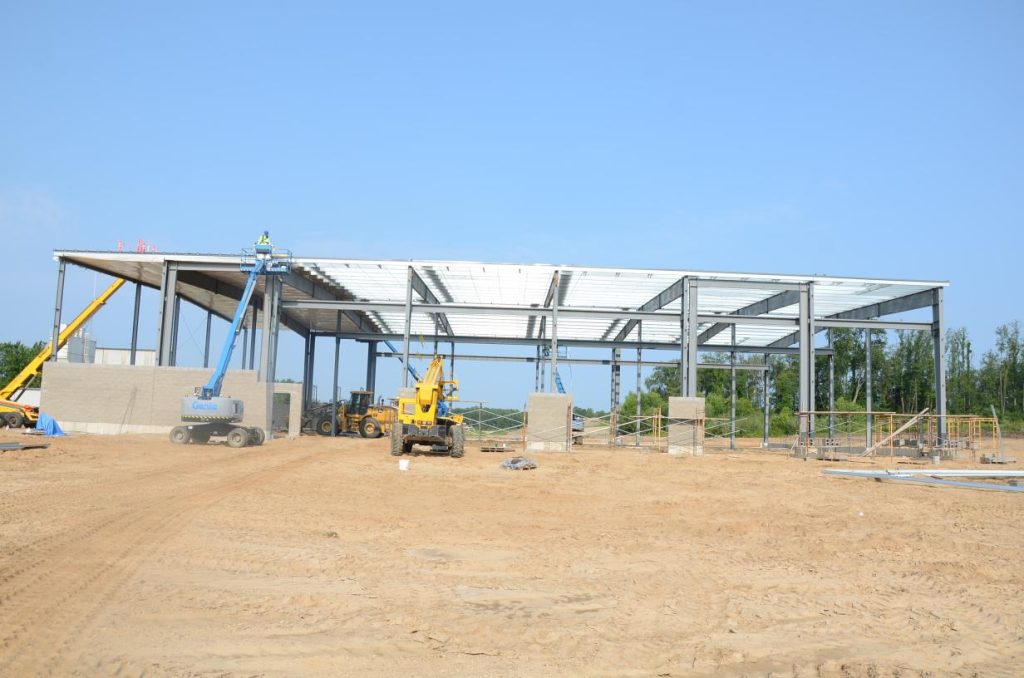 Construction site with a partially built steel frame structure, cranes, and other machinery on a dirt ground.