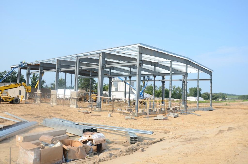 Steel frame structure under construction on a sandy site with scattered materials and a yellow construction vehicle nearby.