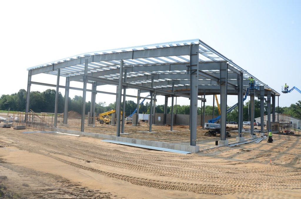 Steel frame construction site with machinery and workers on a sandy ground, surrounded by trees under a clear sky.