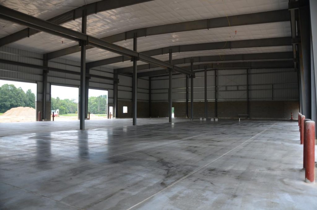 Interior of an empty, large industrial warehouse with high ceilings, steel beams, and concrete floors. Natural light enters through windows and open entrances.