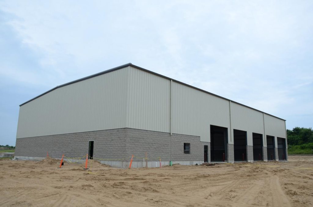 A large industrial building under construction, surrounded by sandy ground and orange safety cones.