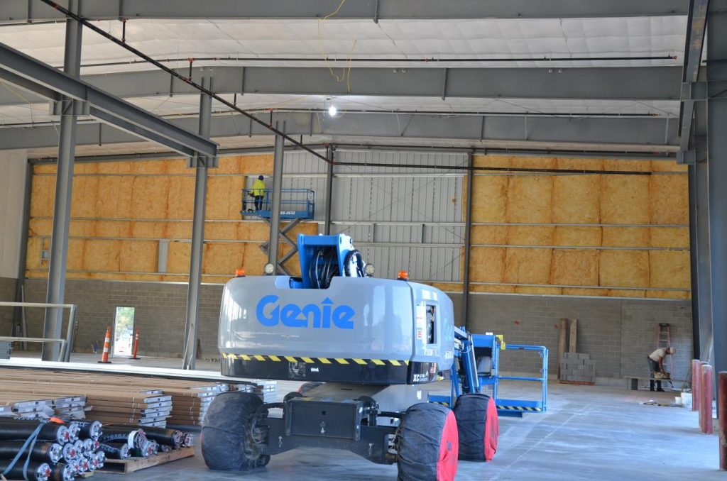 A Genie lift vehicle inside a large industrial warehouse under construction, with insulation on walls and stacks of materials on the floor.