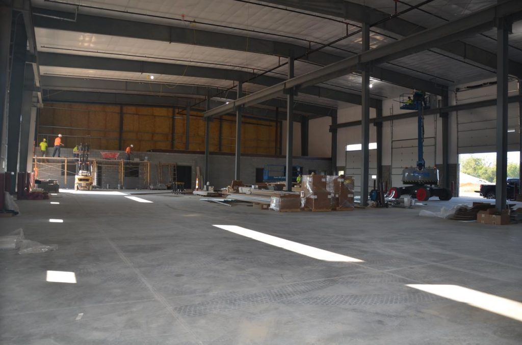 A large, unfinished industrial building with construction workers and equipment inside, including a lift and materials scattered on the floor.