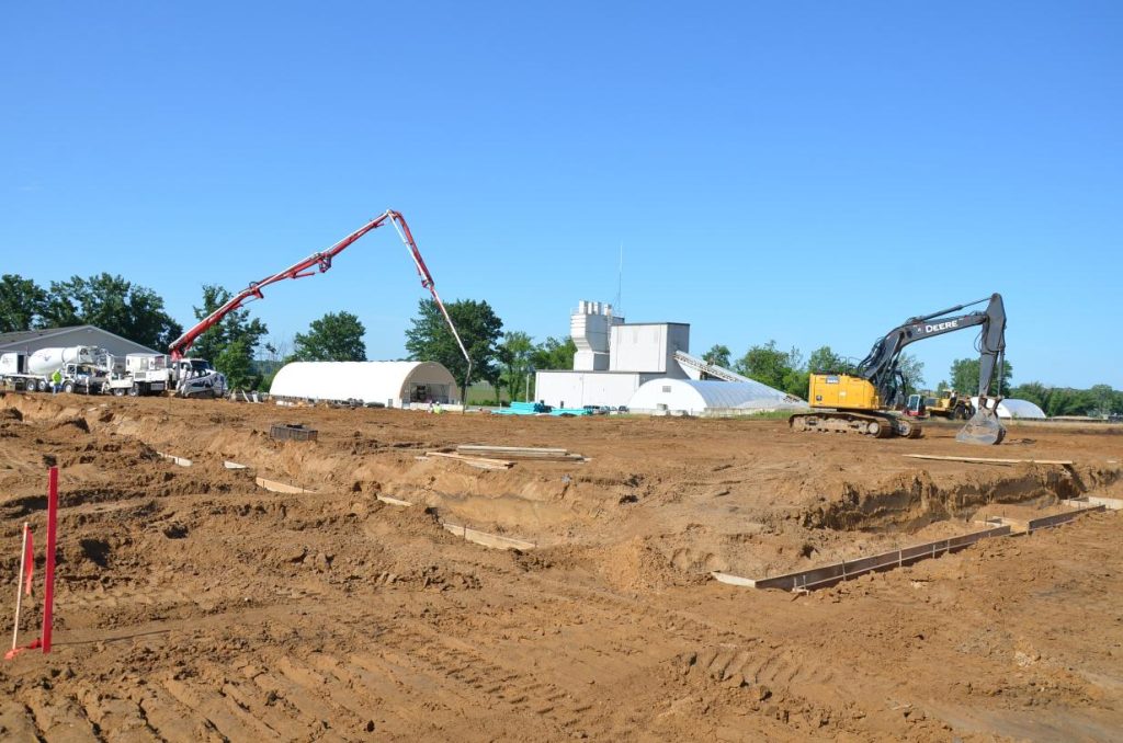 Construction site with an excavator and concrete pump on dirt terrain. Large white tents and trees are in the background under a clear blue sky.