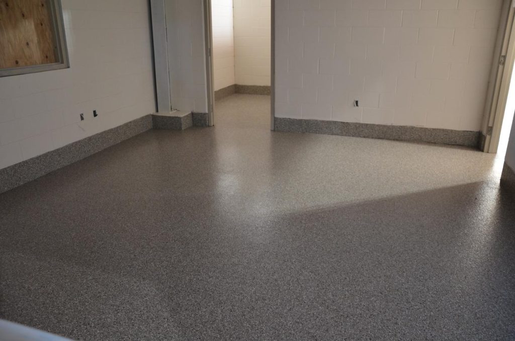Empty room with light gray speckled flooring, white brick walls, and an open doorway leading to another space. Two electrical outlets are visible on one wall.