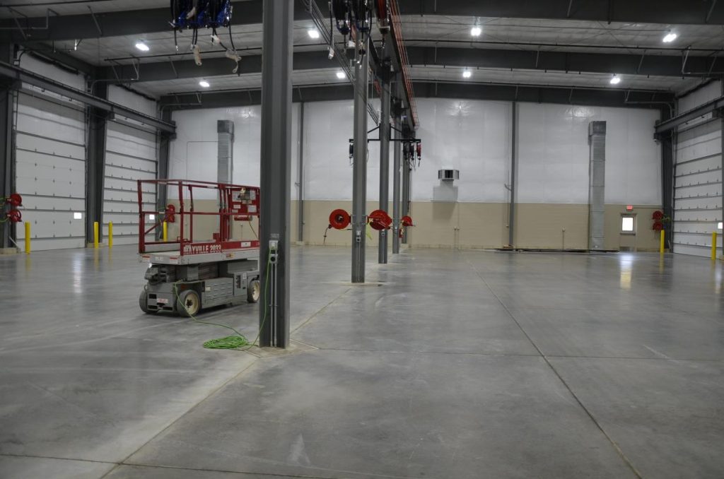 An empty industrial warehouse interior with high ceilings, concrete floors, and multiple doors. A red scissor lift is parked to the left. Ceiling pipes and fire hoses are visible.