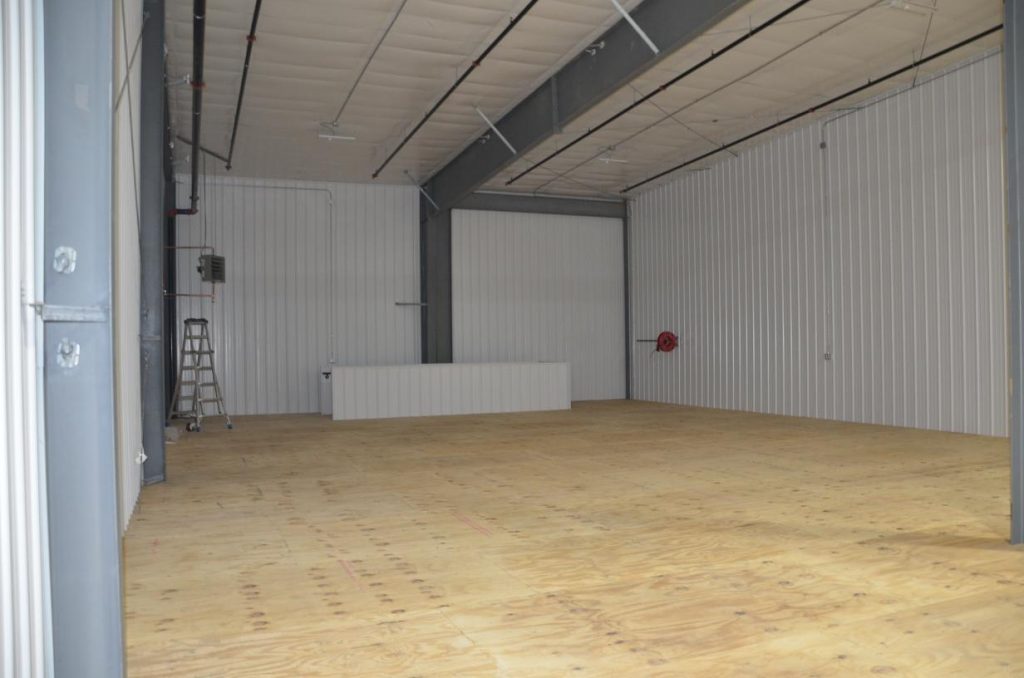 Empty warehouse with wooden floors, white paneled walls, high ceiling, and a ladder in the corner. A desk and red fire hose are against the wall.