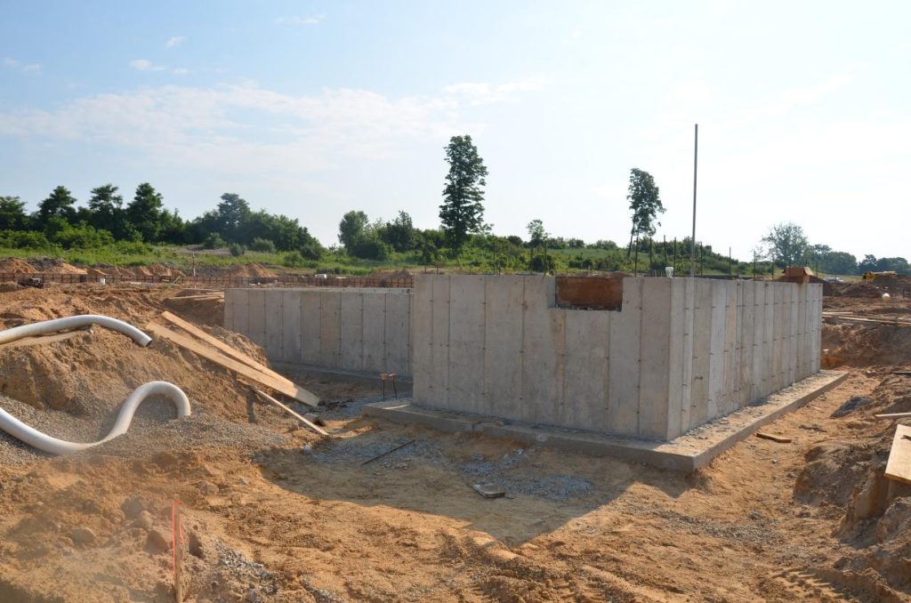 Construction site with partially built concrete foundation walls surrounded by dirt and construction materials. Trees and a clear sky are visible in the background.