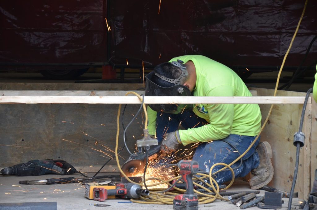 A person in a green shirt and protective gear is using a power tool to cut metal, with sparks flying around. Various tools and cables are scattered on the floor.