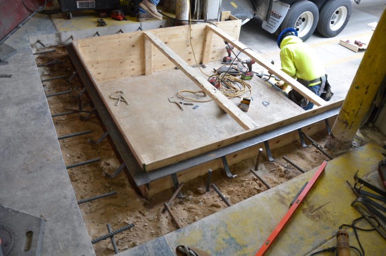 Construction worker installing a concrete loading dock with rebar and wooden framing. Various tools and equipment are scattered around the area.