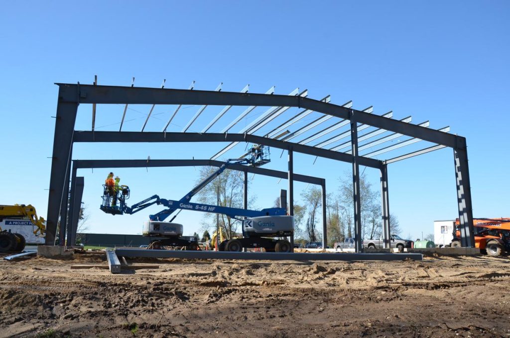 Construction site with steel frame of a building in progress; workers on cranes are installing beams under a clear sky.