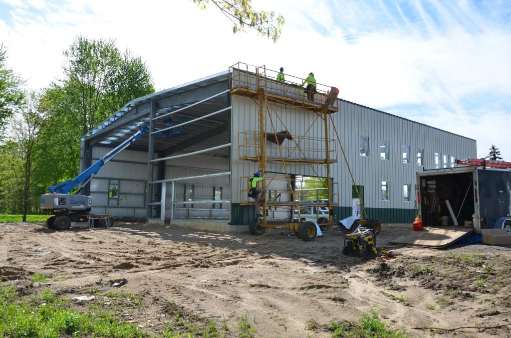 Construction workers building a large industrial structure using scaffolding and a lift, surrounded by trees and construction equipment.