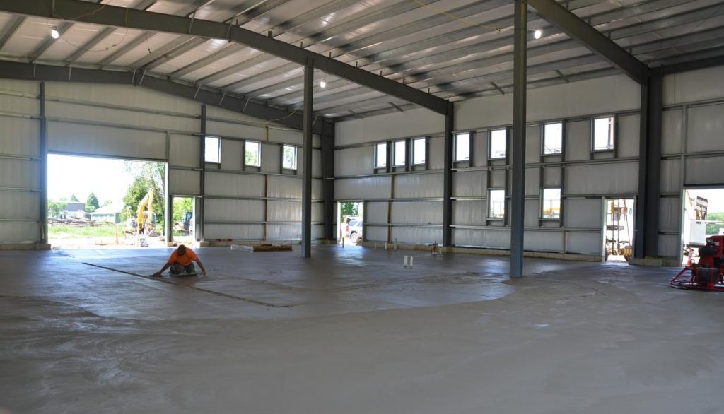 A person works on a large, partially finished concrete floor inside a spacious metal warehouse with high ceilings and multiple windows. Construction equipment is visible outside.
