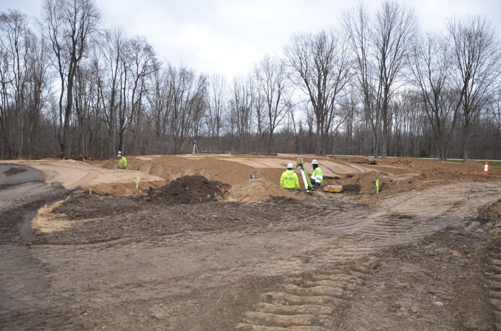 Construction site with several workers in high-visibility jackets and helmets working on a sandy area, surrounded by bare trees under a cloudy sky. Tracks from heavy machinery are visible.