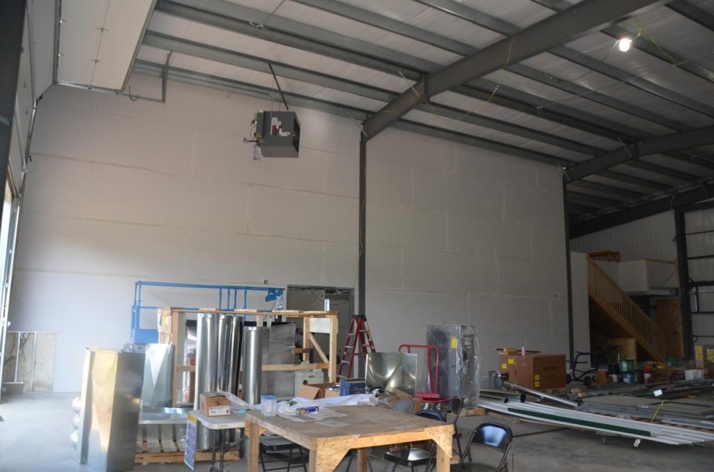Interior of a warehouse under construction with metal beams, scattered materials, ladders, a wooden table with papers and chairs, and an unfinished staircase in the background.