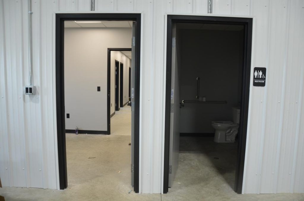 Two open doors in a hallway; left leads to another corridor, right to a restroom with a toilet and grab bar, indicated by a sign. Walls are paneled in white. Floor is concrete.