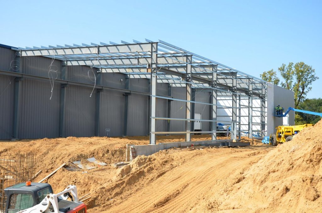 Construction site of a metal warehouse with partially built framework, surrounded by sandy terrain. A small vehicle and workers are visible on site.
