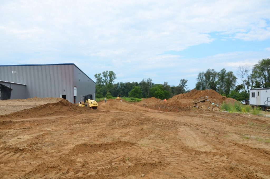 Construction site with a large metal building on the left and piles of dirt. A small construction vehicle is parked in the center. Trees and a cloudy sky are in the background.