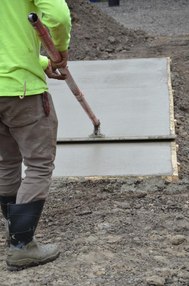 A worker in boots and a green shirt smooths wet concrete on a sidewalk using a long-handled tool.