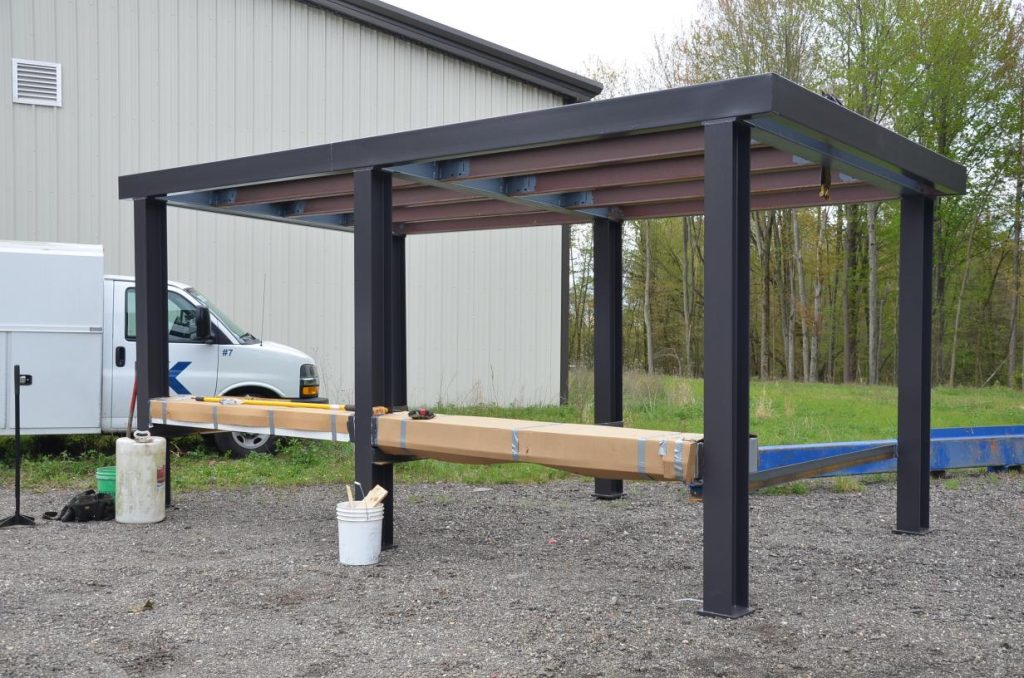 A partially constructed metal canopy structure stands on gravel near a white utility van and a beige building, with construction materials nearby.