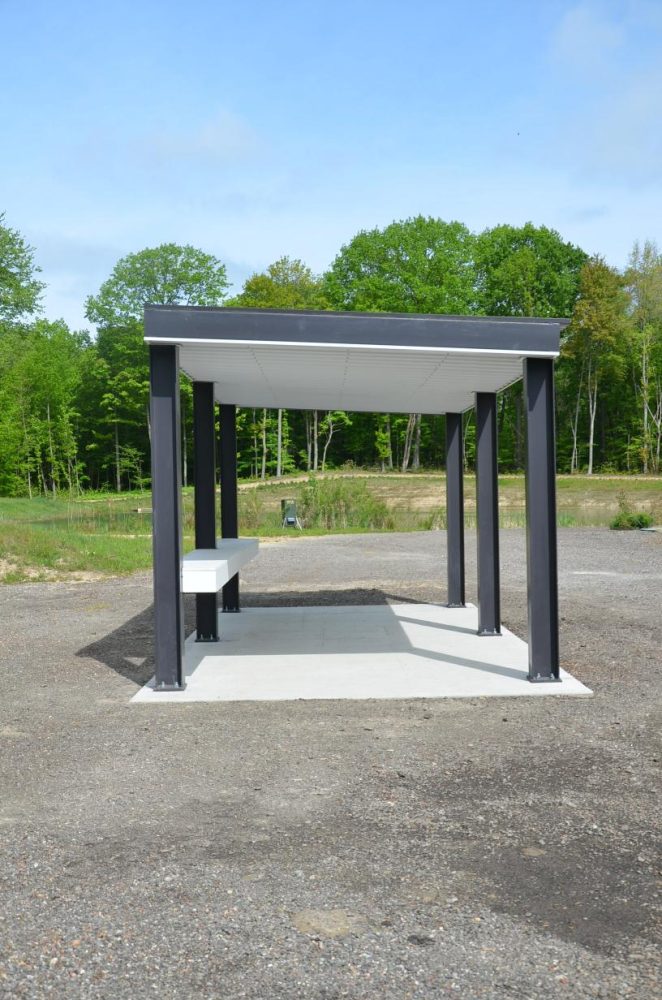 A small, open-sided metal shelter with a flat roof and a bench, situated on a gravel area surrounded by greenery and trees.