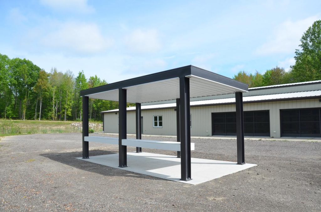 A metal pavilion with a white roof and black supports stands on a concrete pad, in front of a large industrial building with multiple garage doors.