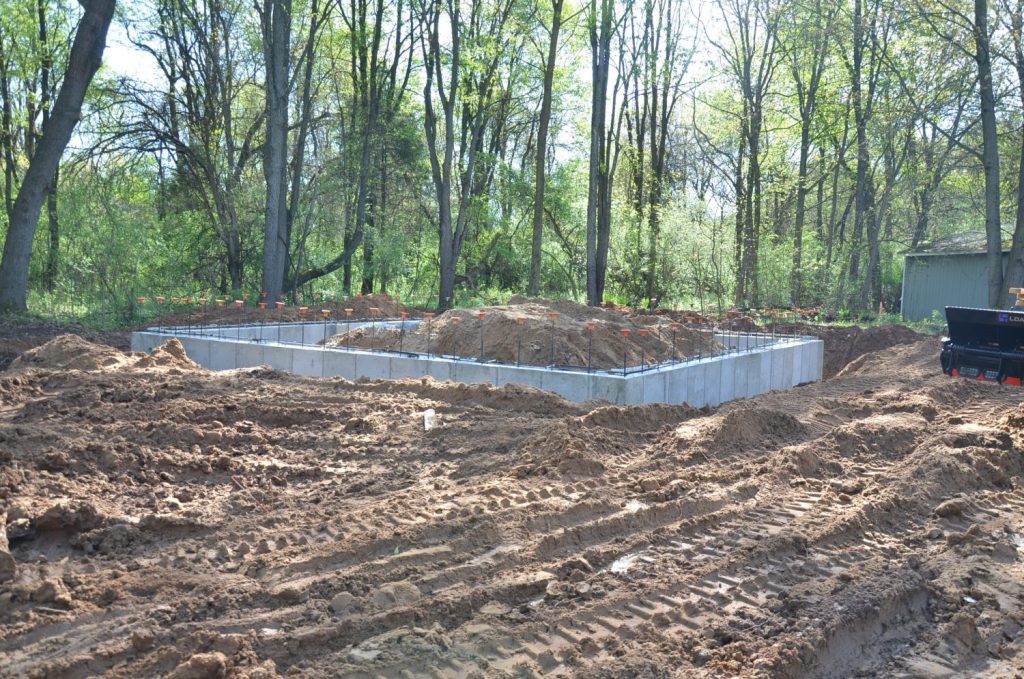 A concrete foundation has been laid at a construction site in a wooded area, with soil and tire tracks visible around the site.