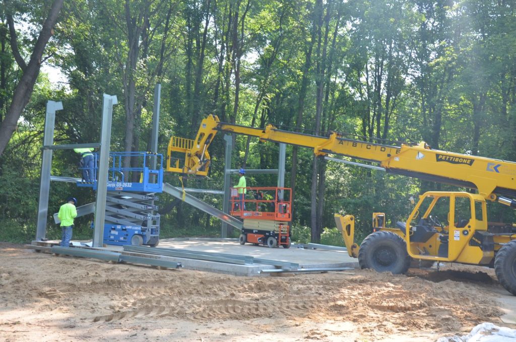 Construction workers use lifts and a yellow telehandler to assemble a steel frame structure on a dirt foundation at a wooded site.