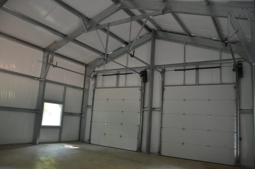 Interior of a metal garage with two white roll-up doors, a small window on the left wall, and unfinished concrete flooring.