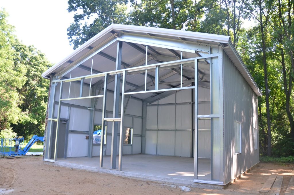 A metal building under construction with exposed steel framing and partially installed siding, set on a concrete foundation and surrounded by trees.