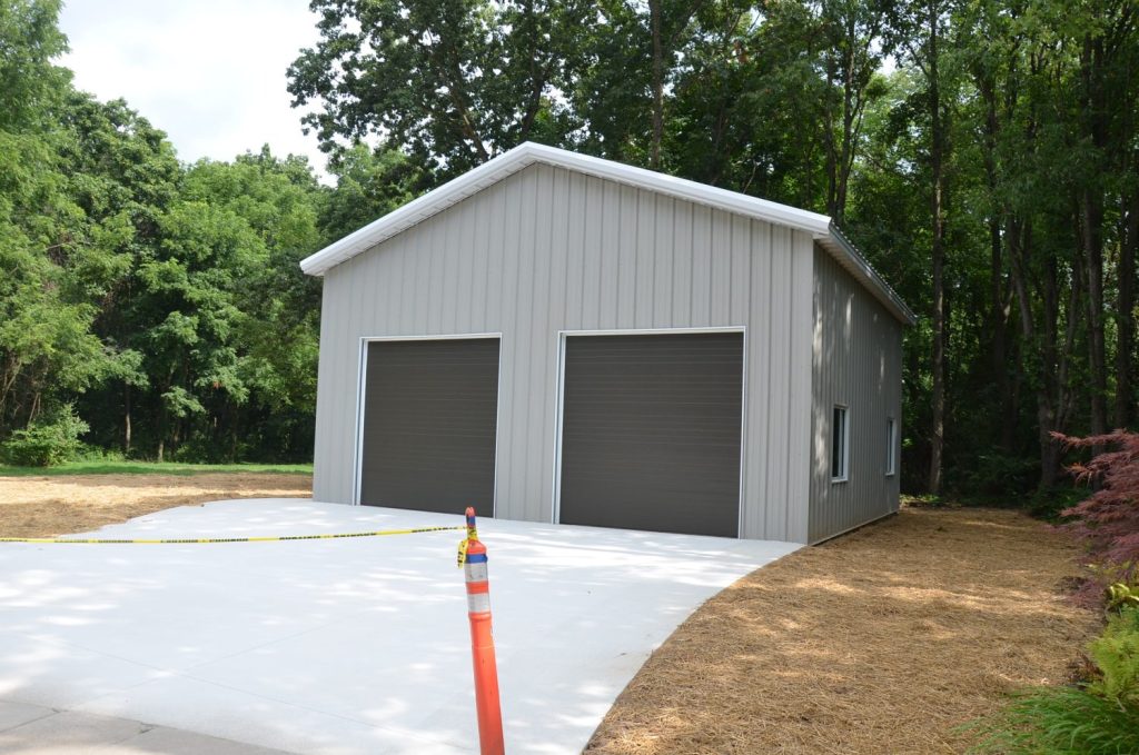 A gray metal garage with two large closed doors sits at the end of a concrete driveway, surrounded by trees; caution tape and an orange traffic cone are in front.