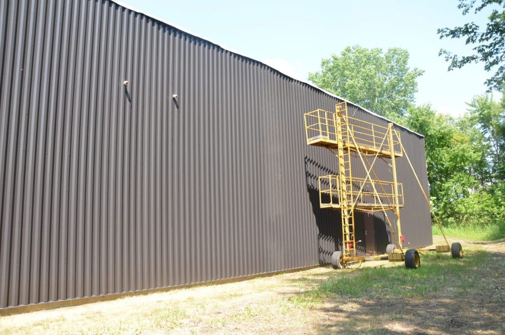 A yellow scaffolding tower is positioned against the side of a large, dark corrugated metal building surrounded by grass and trees.