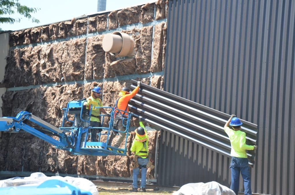Four construction workers, wearing safety gear, install metal siding panels on a building exterior; two use a lift while two work from the ground.