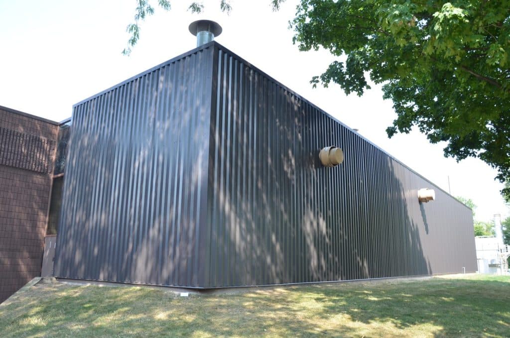 A large, rectangular industrial building with dark corrugated metal siding, ventilation pipes, and a nearby tree providing partial shade.