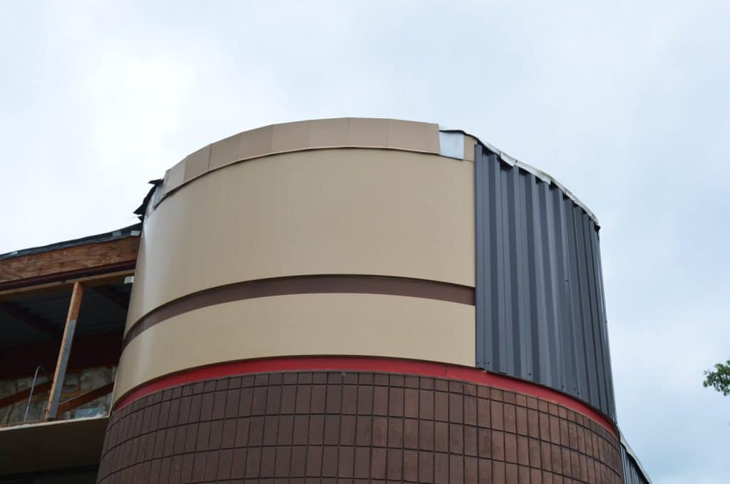 A close-up of a building corner showing unfinished construction with different exterior materials, including brown brick, beige panels, and gray corrugated metal.
