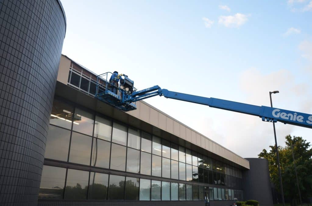 Two workers on a blue boom lift perform maintenance or installation on the exterior upper facade of a commercial building with large windows.