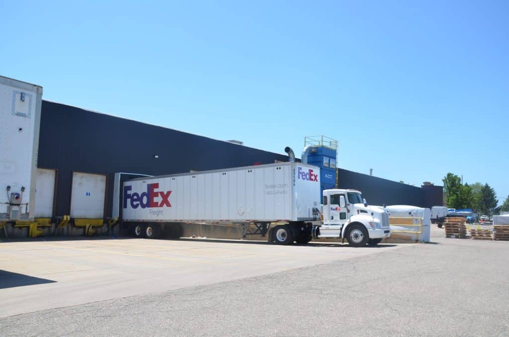 A FedEx Freight truck is backed up to a loading dock at an industrial warehouse on a clear, sunny day.