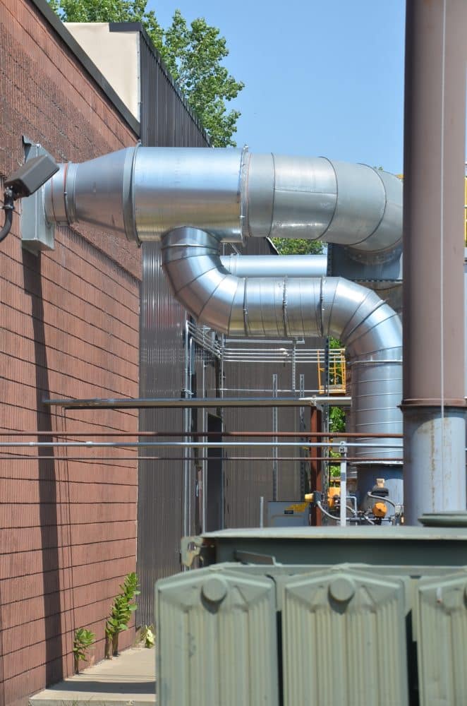 Large metal ventilation ducts run along the exterior of a brick industrial building, with some small plants growing at the base.