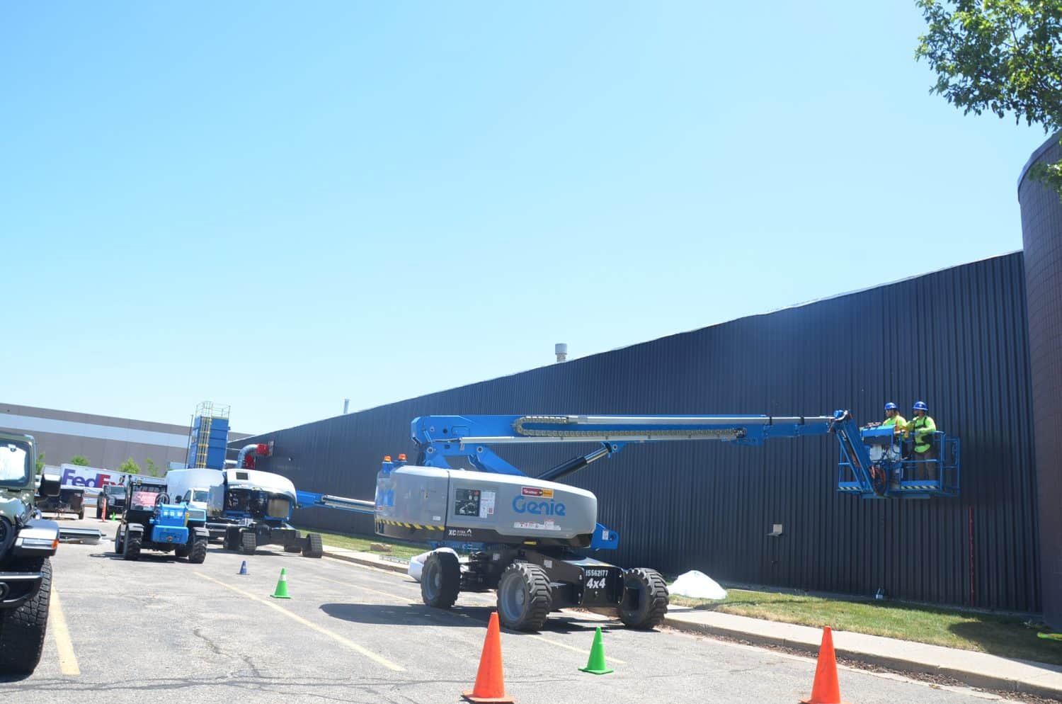Workers in safety gear operate a Genie boom lift to access the side of a large industrial building, with traffic cones marking the work area.