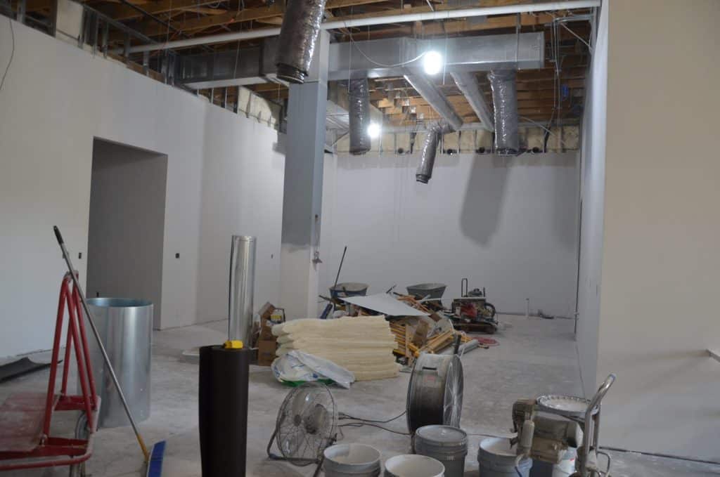 Interior of a building under construction with unfinished walls, exposed ceiling ducts, construction materials, fans, and tools scattered on the floor.