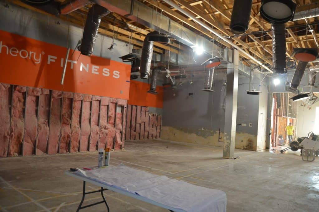 Interior of a fitness studio under construction, with exposed insulation, ceiling ducts, construction materials, and a table covered with a white cloth in the foreground.