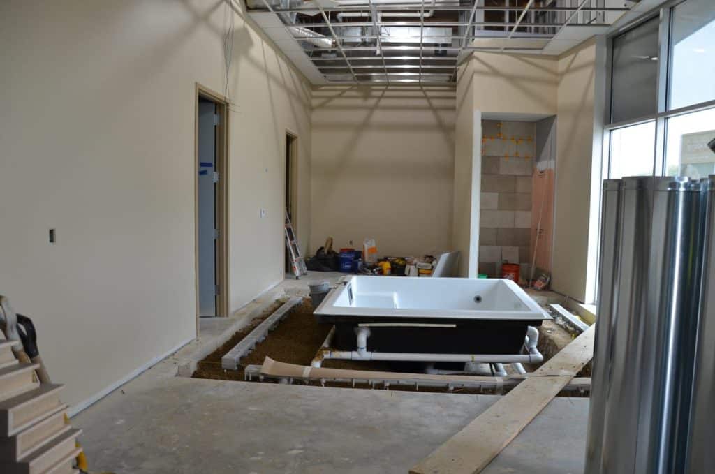 A construction site shows a partially installed bathtub in the ground, exposed pipes, tools, and building materials in an unfinished room with visible ceiling framework.