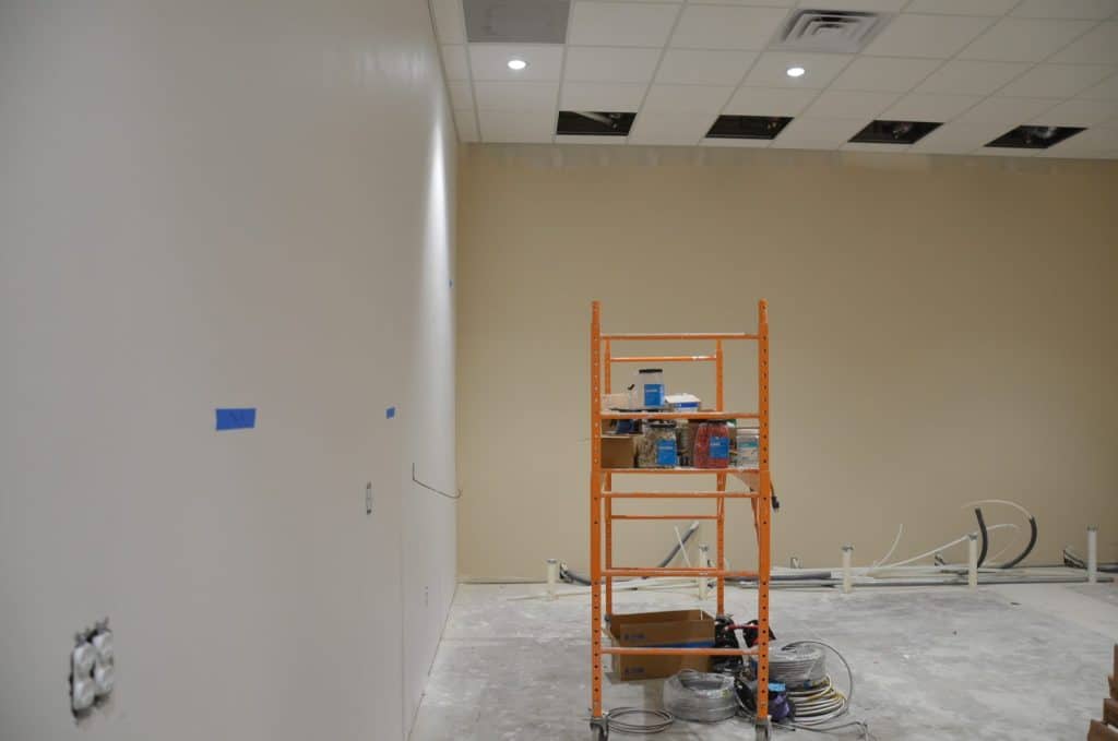 A partially finished room with exposed ceiling tiles, a scaffold holding painting supplies, loose wires on the floor, and taped spots on the wall, indicating ongoing construction.