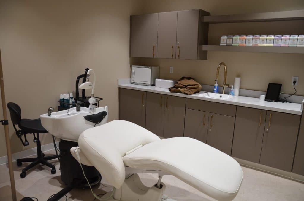 A modern treatment room with a reclining chair, a hair washing sink, cabinets, and shelves with beauty products and supplies.