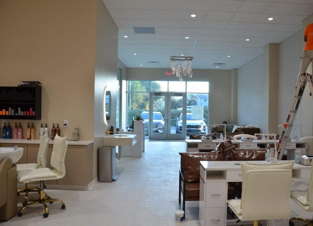 A modern nail salon interior with manicure stations, white chairs, beauty products on shelves, and a worker on a ladder near the ceiling. Large windows show parked cars outside.